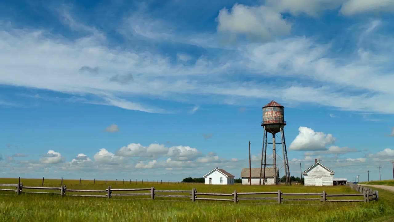 A weathered wooden water tower at the edge of a small rural American town under a bright noon sky, faded painted markings on the tank.
